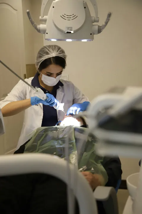 Dentist working on a patient in a dental office. Both wear masks. Bright light illuminates.